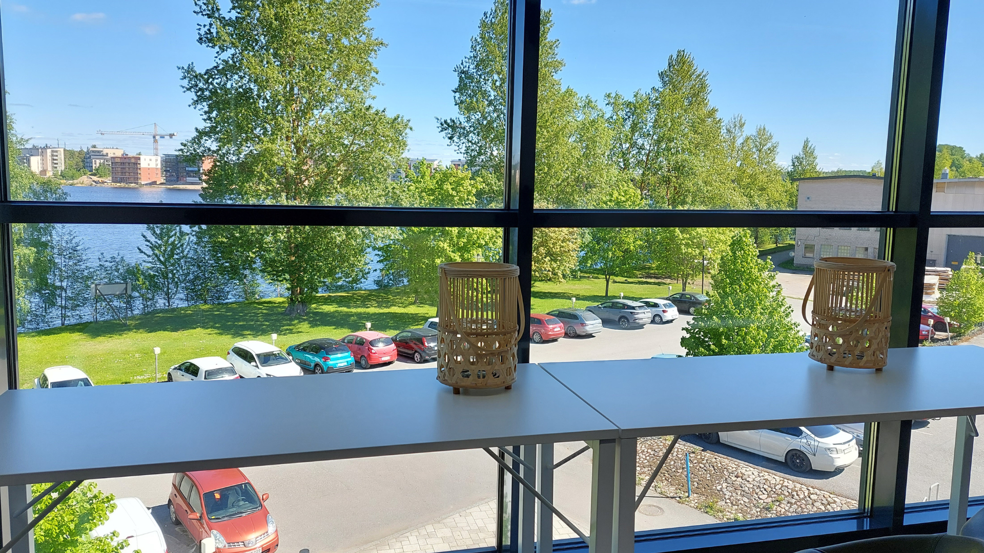 Tässä kuvassa on Meitan toimistolta otetun maisematiskin pöytää, kaksi kynttilälyhtyä ja näkymä ikkunasta ulos kesäiseen maisemaan