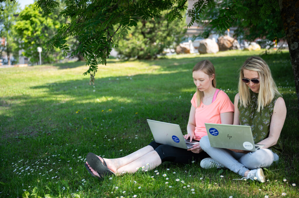 Tässä kuvassa kaksi meitalaista istuu ulkona nurmikolla, keskittyneenä työskentelemään kannettavat sylissään